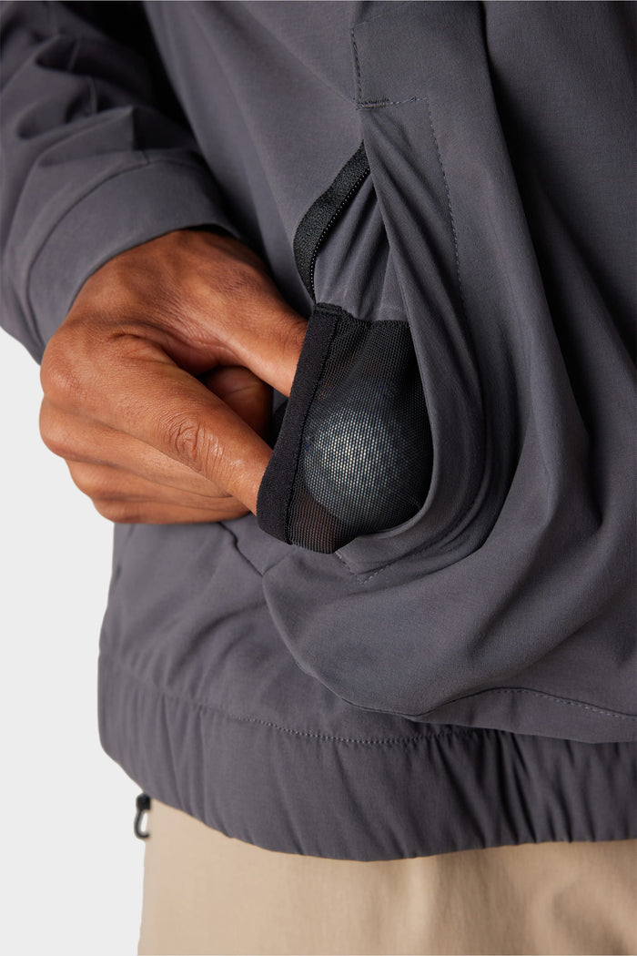 A hand places a silver ball into the black mesh pocket of the 686 x Malbon Men's PrimaLoft drirelease® Merino-Lined Pullover Jacket, with part of the gray jacket and beige pants visible.