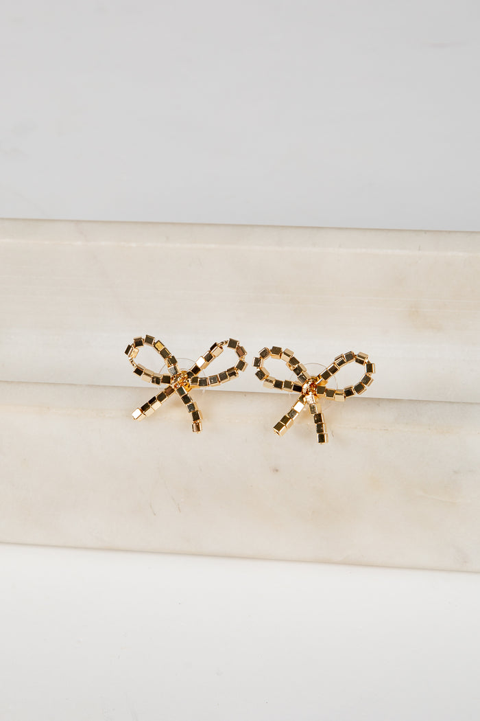 Gold bow-shaped earrings on a beige surface with a light gray background