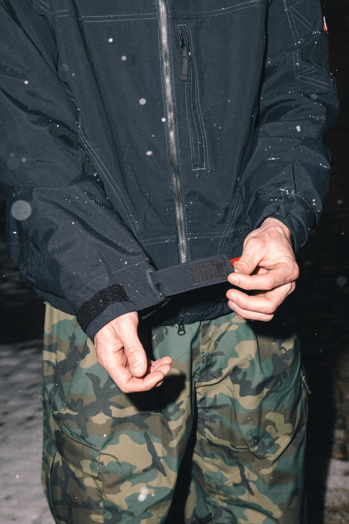 A person wearing the 686 Men’s Dojo® Shell Jacket adjusts the strap on their sleeve, while snowflakes fall around them.