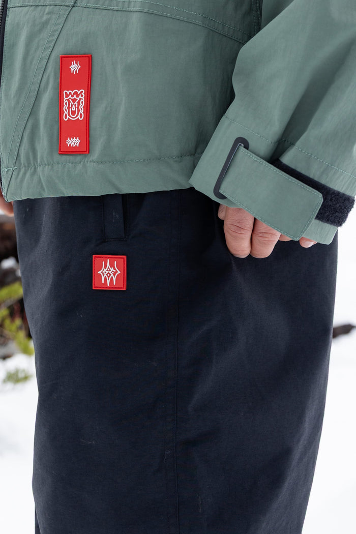 A close-up of a person wearing a green jacket and black 686 Men’s Dojo® Shell Pants, both featuring red rectangular patches with white graphic designs, standing outdoors in a snowy environment.