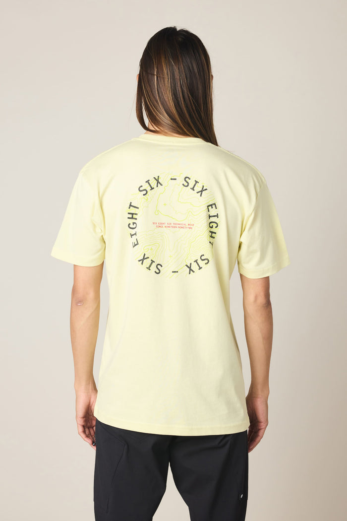A person with long hair faces away from the camera, wearing the 686 Lifts Authentic SS Tee‚Äîa light yellow 100% cotton T-shirt by 686 featuring a circular ‚ÄúSIX EIGHT SIX‚Äù graphic and text on the back, set against a neutral background.
