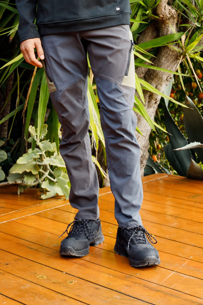 A person stands on a wooden deck wearing 686 Men's Anything Cargo Pant - Slim Fit in gray and black hiking boots, surrounded by green plants‚Äîready for adventure.