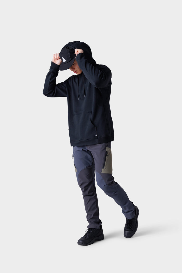 Wearing a black hoodie, black cap, and 686 Men's Anything Cargo Pant - Slim Fit by 686, paired with black sneakers, a person stands against a plain light gray background, adjusting the cap‚Äîready for any adventure.
