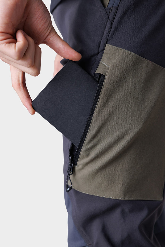 A person‚Äôs hand holds a black card partly inside the zippered pocket of 686 Men's Anything Cargo Pant - Slim Fit by 686, shown in black and olive green color-blocked design against a plain background.