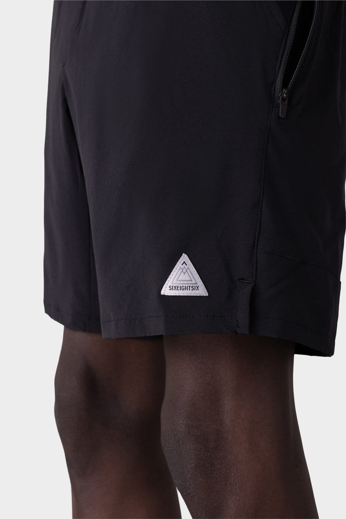 A close-up of a person wearing 686 Mens ATP Stretch Performance Shorts shows the black fabric, small white triangular 686 logo patch near the hem, part of their upper legs, and the shorts zipper pocket.