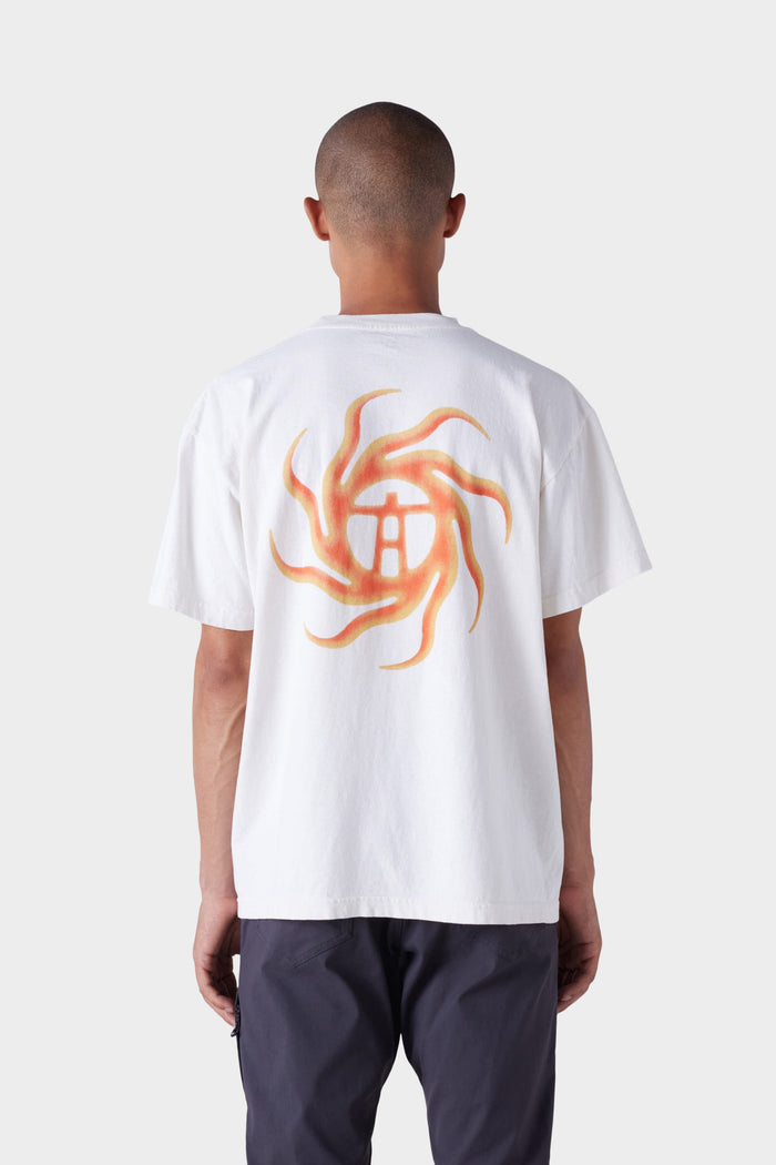 A person with short hair wears dark pants and the 686 Mens Man in the Sun Short Sleeve Tee by 686, featuring a white color with an orange and yellow swirling sun design and central symbol on the back.