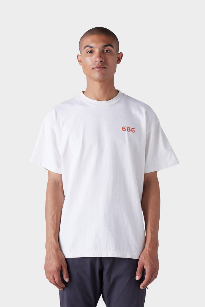 A person stands facing forward, wearing the 686 Mens Man in the Sun Short Sleeve Tee‚Äîa loose white T-shirt with small red 686 text on the upper left chest‚Äîpaired with dark pants, set against a plain light background.