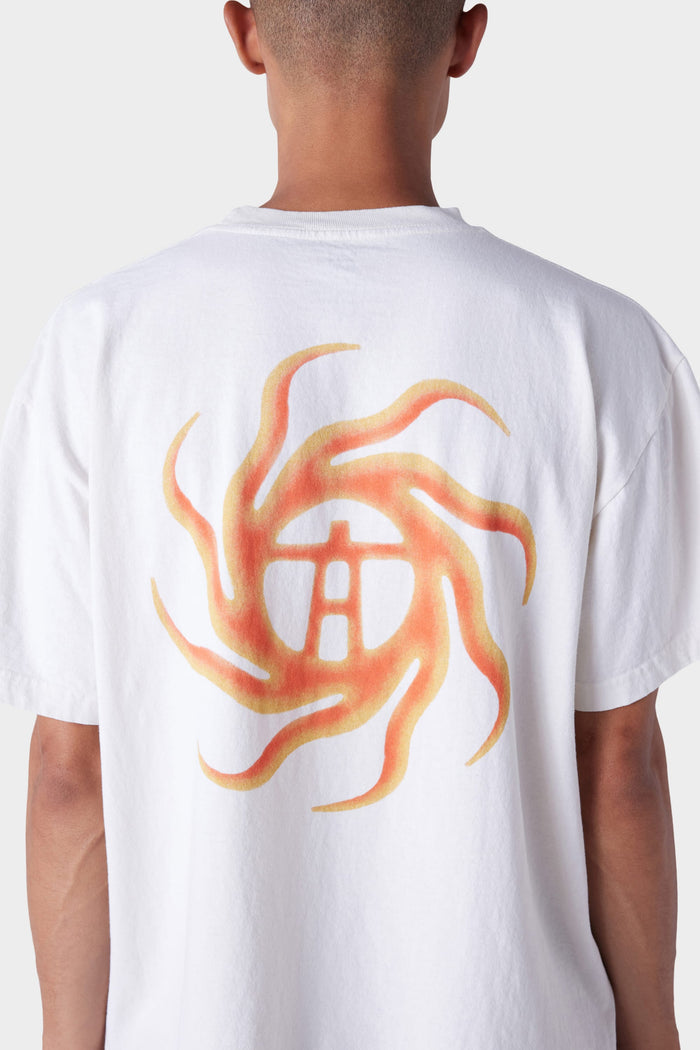 A person wears the 686 Mens Man in the Sun Short Sleeve Tee by 686, featuring a white base and an orange-yellow swirling sun design with a central symbol on the back, standing against a plain light background.