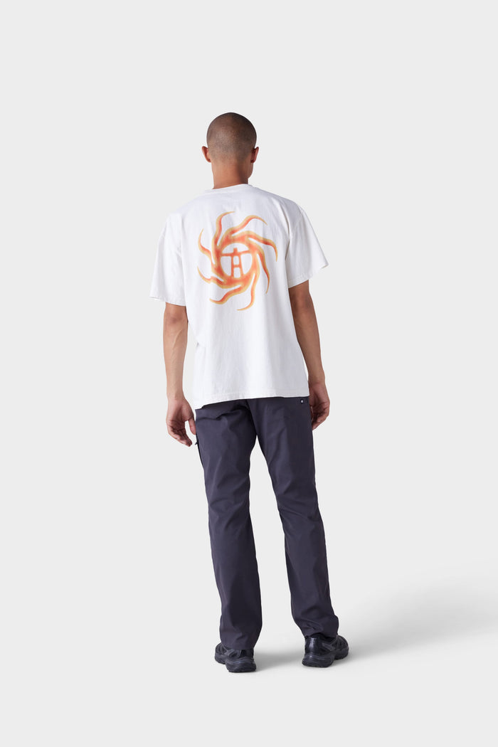 A person faces away, wearing the 686 Mens Man in the Sun Short Sleeve Tee by 686 with an orange abstract sun and globe design on the back, paired with dark pants and black shoes against a plain light gray background.