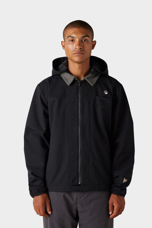 A person faces forward against a light gray background, wearing the 686 x Malbon Mens Caddie Performance Jacket by 686‚Äîa black hooded jacket with a front zipper and a small circular logo on the left chest.