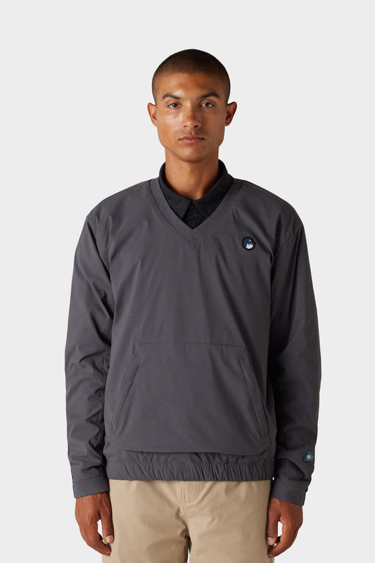 A young man with short hair and a neutral expression wears the 686 x Malbon Men's PrimaLoft drirelease® Merino-Lined Pullover Jacket in dark gray, featuring a small logo on the left chest, paired with khaki pants against a light background.