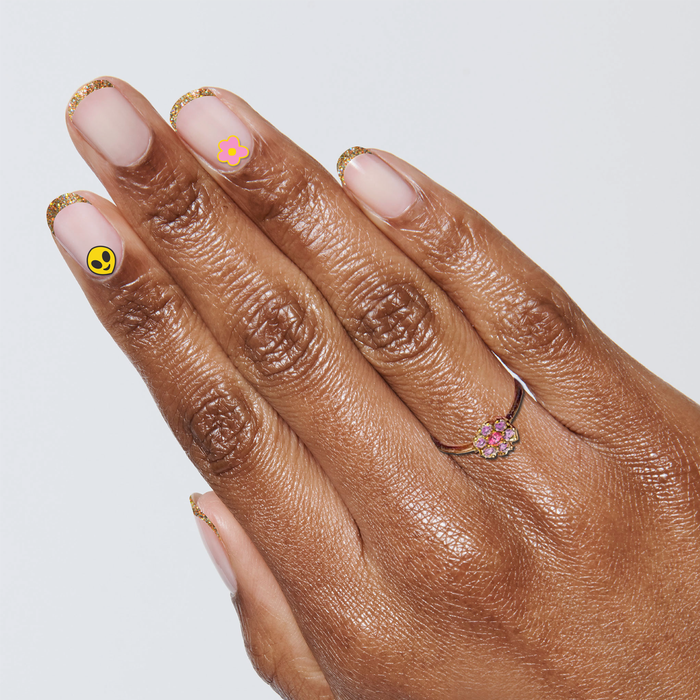 Hand with neutral polish edged in gold glitter and Frasier Sterling Pack stickers of a pink flower and yellow alien emoji for manicure