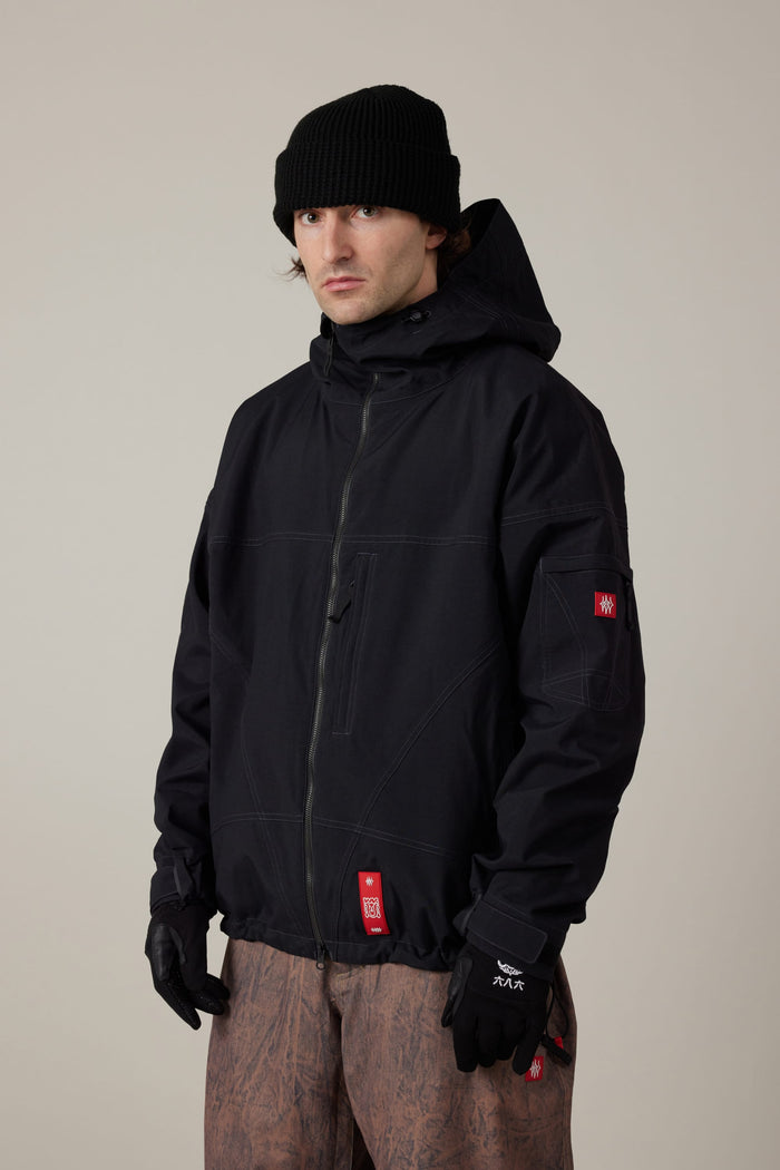 A person wearing the 686 Men’s Dojo® Shell Jacket in black stands against a plain background, paired with a black knit beanie and black gloves. The jacket, designed in the style of a men’s woven jacket with visible stitching and red tags, features a hood and is made from polyester. The individual has a neutral expression and looks slightly to the side.