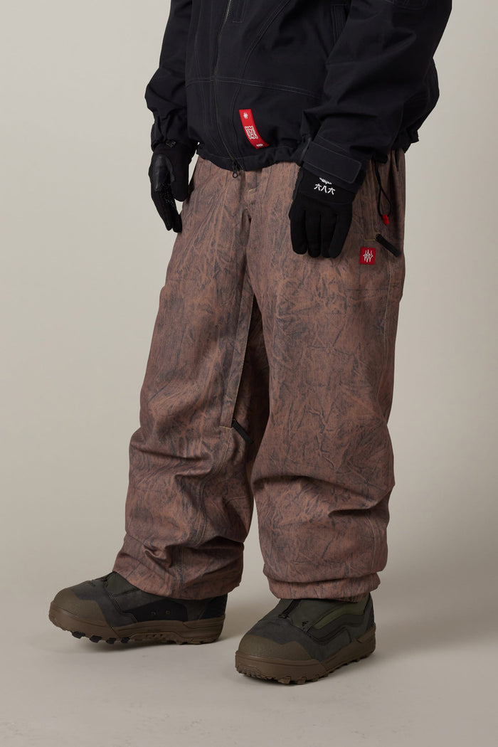 A person is wearing 686 Men’s Dojo® Shell Pants—baggy, brown, and patterned waterproof pants—along with dark gloves, a dark jacket, and rugged green snow boots, standing against a plain background.