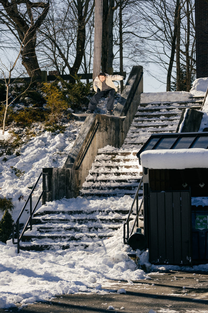 Wearing a white jacket and the Men’s 686 Dojo® Shell Pant by 686, a snowboarder slides down a concrete handrail beside snowy outdoor steps, surrounded by sunlit snow-covered ground and trees.