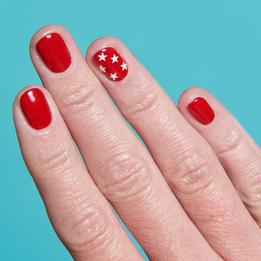 Hand with bright red polish featuring white star nail stickers on the ring finger for Stars mani on blue background