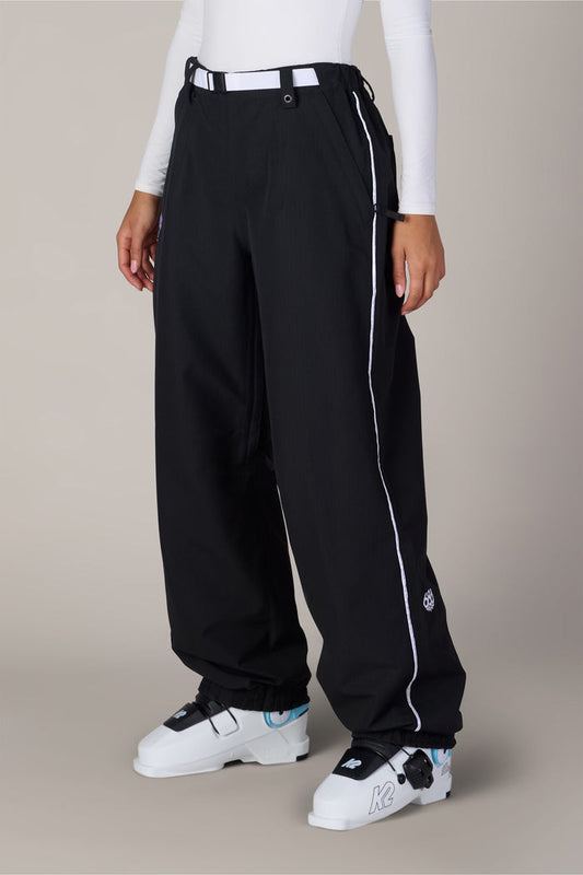 A person wears the 686 Women’s Hellkat™ Shell Pant in black with white piping, paired with a white long-sleeve top and white ski boots, standing against a plain background.