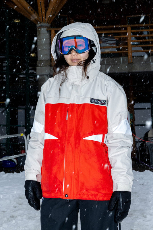 A person stands outdoors in falling snow, with a building and snow-covered ground in the background, wearing the 686 Women’s Search Jacket paired with black gloves and ski goggles.